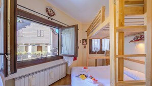 A bedroom with a wooden bunk bed, a window with curtains, a radiator, and a small stool.