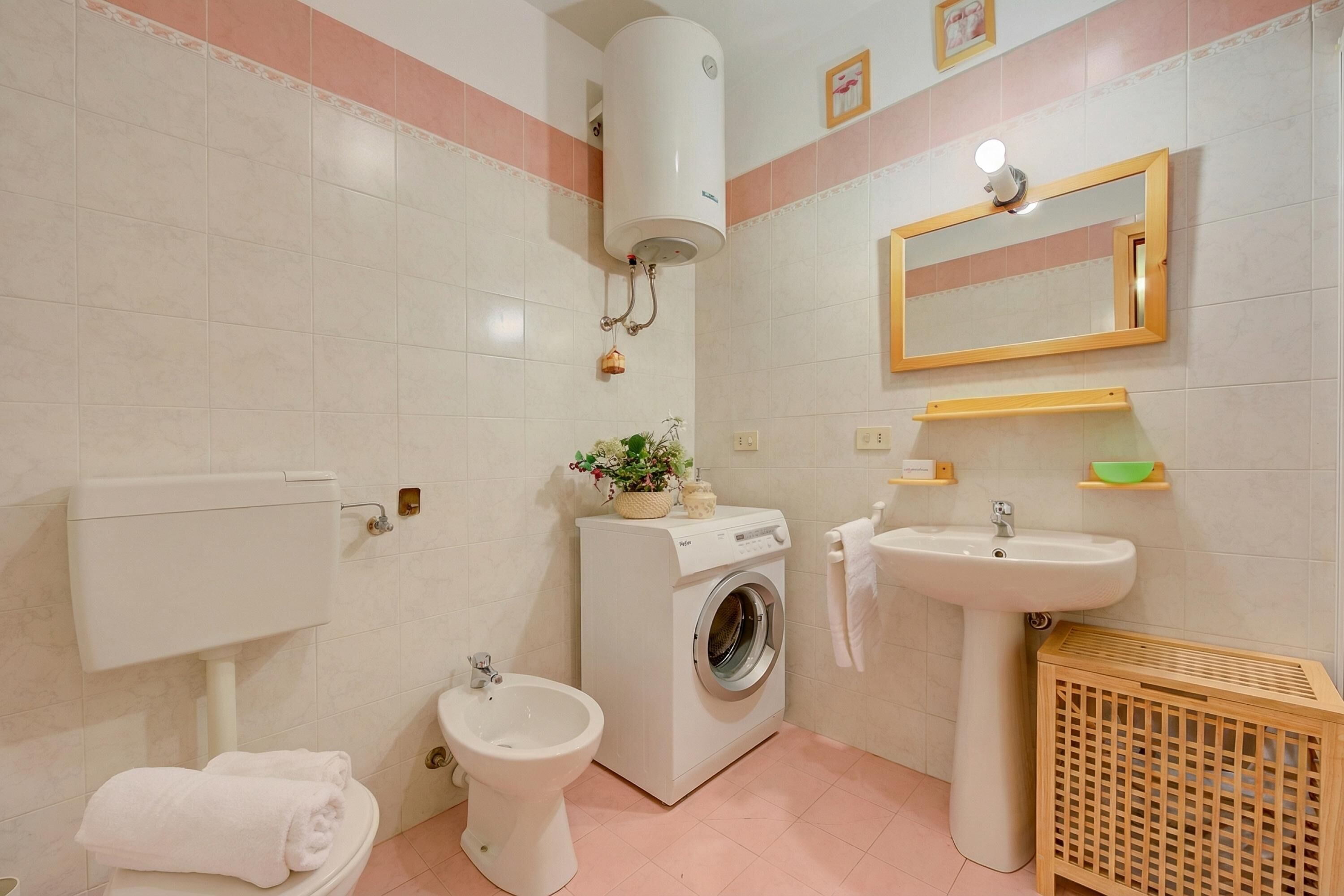 A bathroom with a toilet, bidet, washing machine, sink, mirror, and a wooden basket.