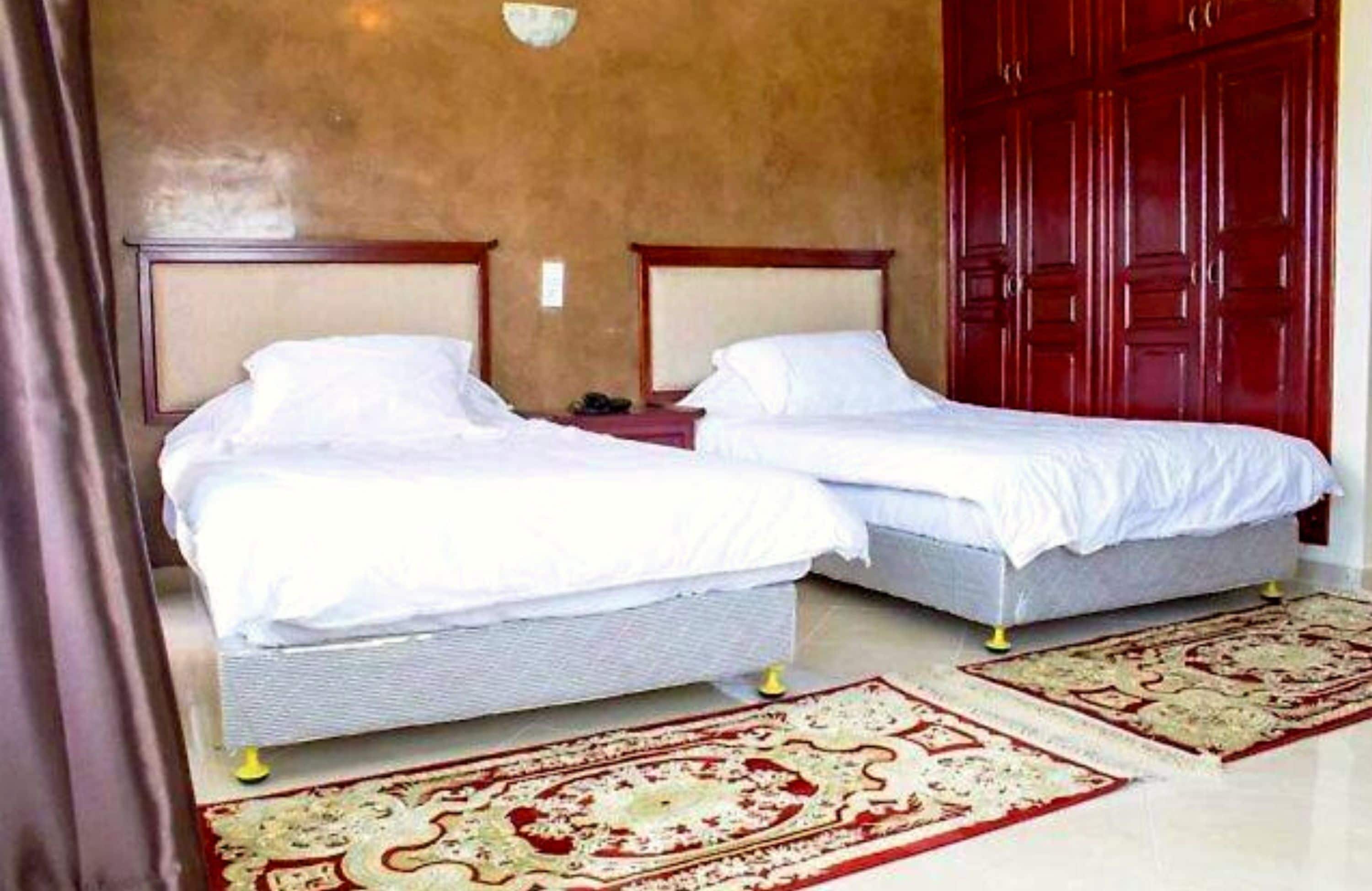A hotel room with two single beds, a wooden wardrobe, and a red and beige patterned rug.