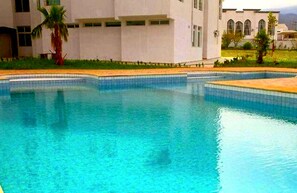 A swimming pool with clear blue water, surrounded by a white building with large windows and a palm tree. - Angle Atlas Hotel (Dir El Ksiba)