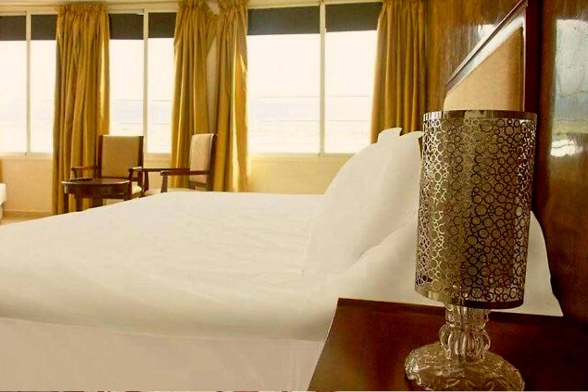 A hotel room with a large bed, a bedside lamp, and a view of the sea through the windows.