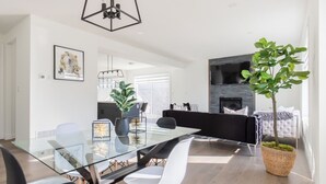 A modern living room with a glass dining table, black and white chairs, a black sofa, a fireplace, and a potted plant. - Comfortable Lux | Sleeps 10 | PingPong, Cozy Family Movie Room (London)