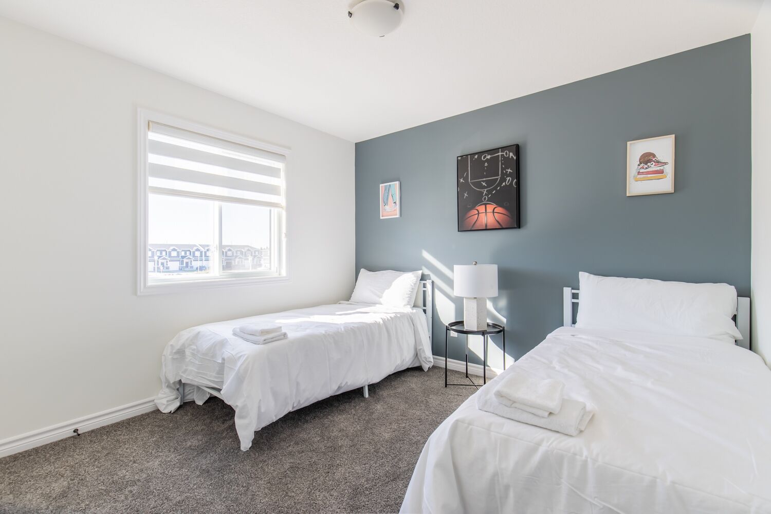 A bedroom with two beds, a window with blinds, a lamp on a side table, and artwork on the walls.