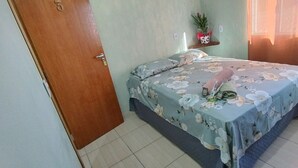 A bedroom with a bed, a wooden door, a small shelf with a plant, and a window with curtains. - Pousada e Parada Colonial (Capitão Leônidas Marques)