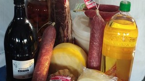 A variety of food items including a bottle of wine, a jar of tomatoes, a large piece of meat, a sausage, a cheese, and a bottle of oil on a checkered tablecloth. - Pousada e Parada Colonial (Capitão Leônidas Marques)