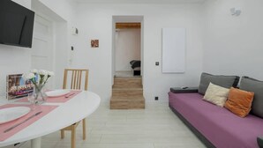 Een moderne woonkamer met een witte eettafel gedekt voor twee personen, een paarse bank met kussens, en een trap die naar een andere kamer leidt.