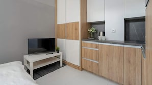 Camera d'albergo moderna con un letto, una TV su un mobile bianco, un tavolino con una pianta e un angolo cottura con pensili in legno e un piano di lavoro in marmo.