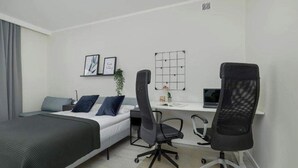 A bedroom with a bed, two office chairs, a desk, a small plant, and a wall-mounted light fixture.