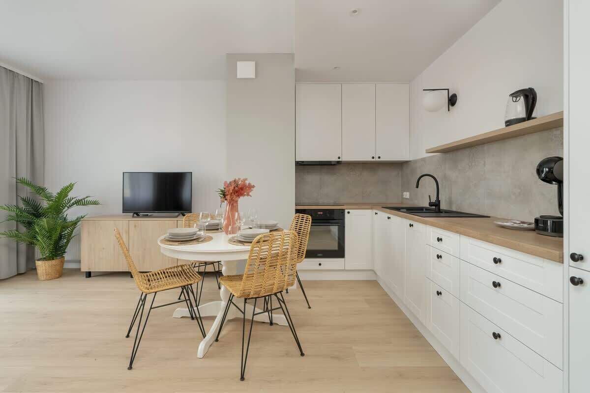 A modern kitchen with white cabinets, a wooden countertop, and a built-in oven. There is a dining area with a white table and wicker chairs.