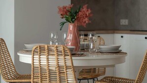 Mesa de jantar redonda com pratos brancos, copos transparentes e um vaso rosa com flores. Cadeiras de fibra natural estão dispostas ao redor da mesa.