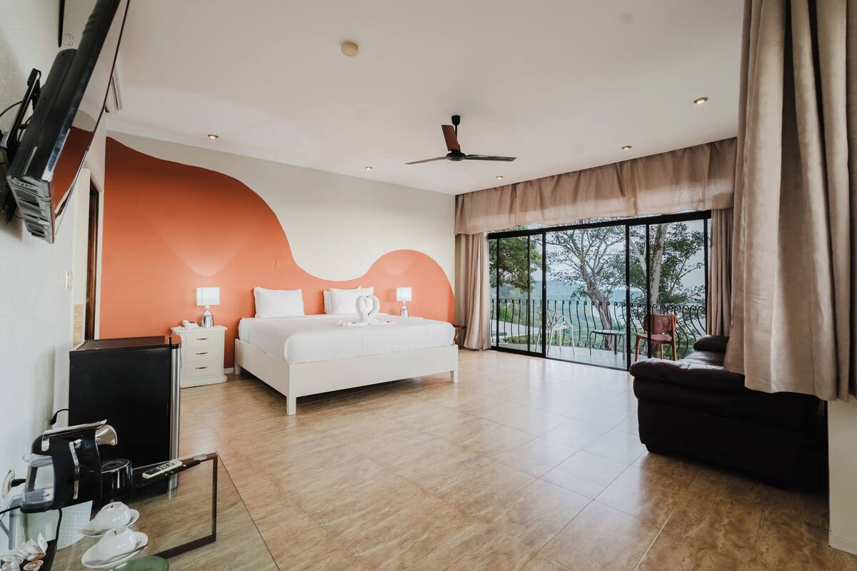 A modern hotel room with a large bed, a flat-screen TV, a desk with a coffee machine, and a view of the outdoors through large windows.