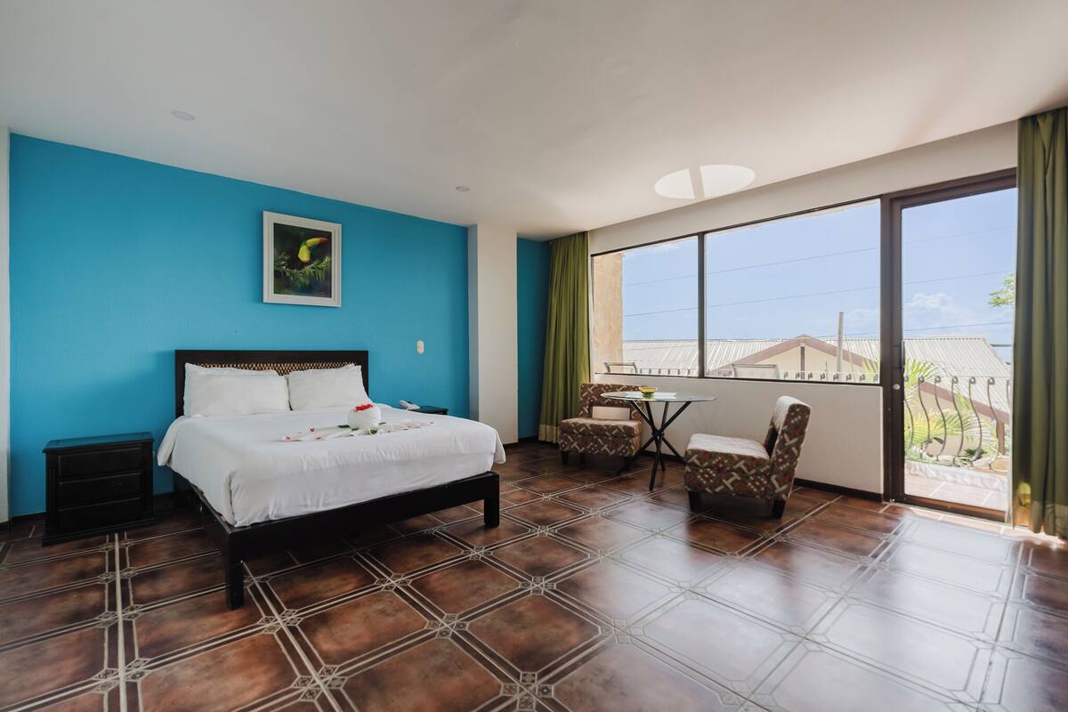 A bedroom with a bed, a small table, two chairs, a nightstand, and a large window with a view of buildings.