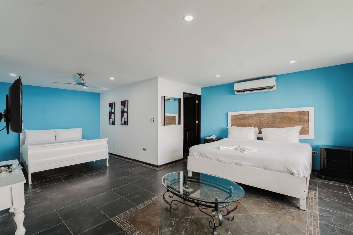 A modern hotel room with a large bed, a glass coffee table, a white dresser, and a ceiling fan.