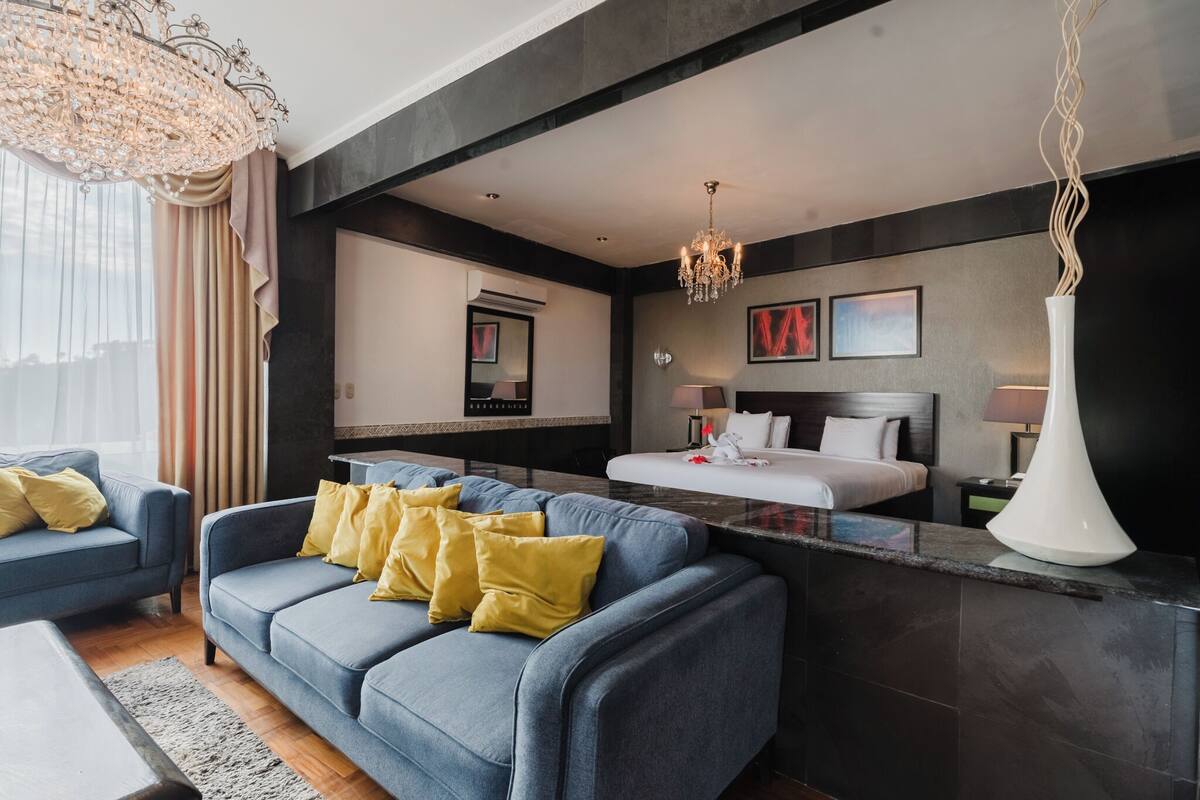 A modern hotel room with a blue sofa, a bed, a large mirror, and a chandelier.