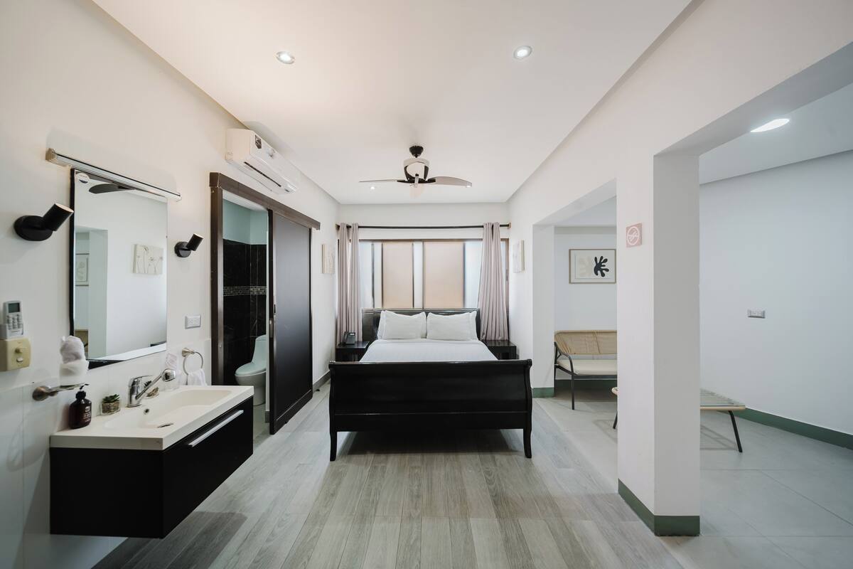 A modern hotel room with a large bed, a bathroom with a vanity, a ceiling fan, and a large window with curtains.