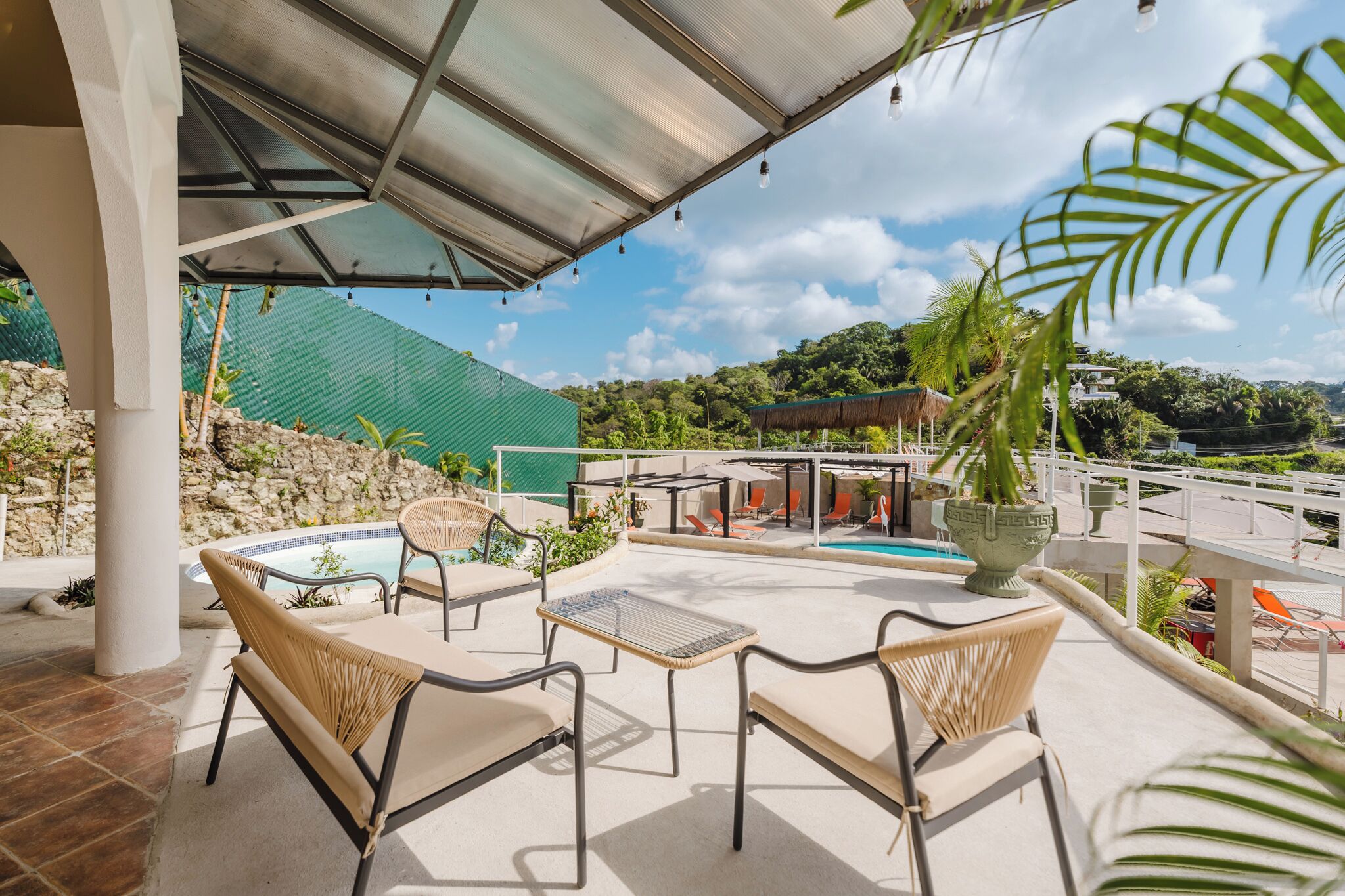 Un espacio de estar al aire libre cubierto con mobiliario de mimbre, una piscina y vistas a un edificio verde y árboles.