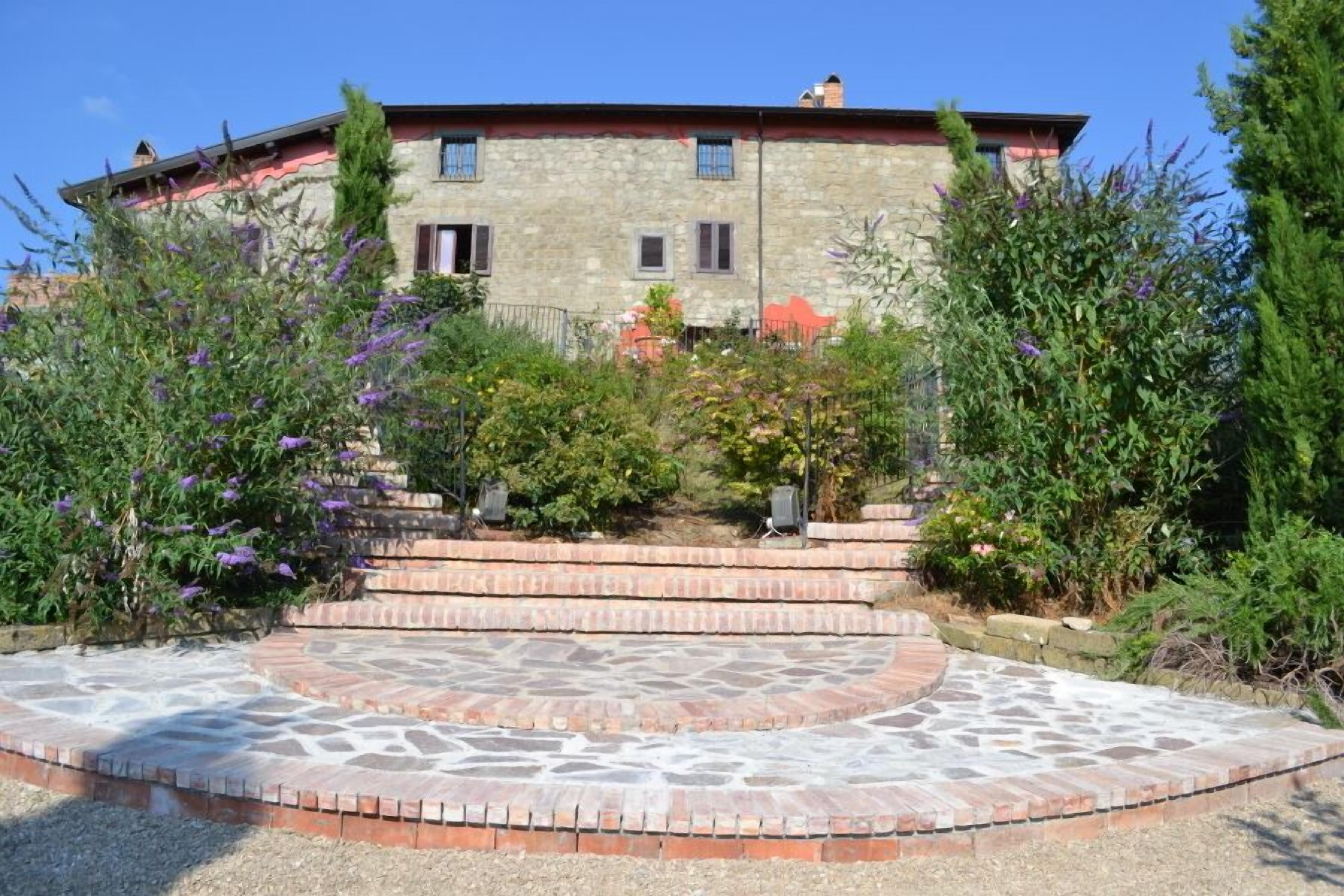 A stone building with a red roof, a paved courtyard with a circular pattern, and surrounding greenery.