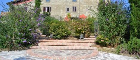 A stone building with a red roof, a paved courtyard with a circular pattern, and surrounding greenery.