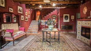 Habitación tradicional con escalera de madera, sofá con cojines, mesa con un florero y chimenea con azulejos decorativos.
