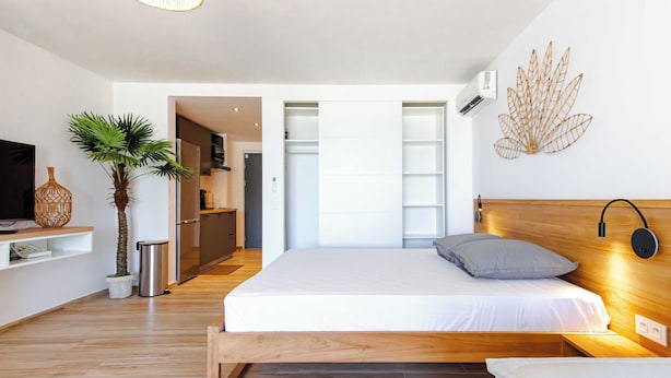A modern bedroom with a wooden bed, a TV, a plant, and a kitchen in the background. - Beautiful studio on the beachfront Baie Orientale (Saint-Martin)