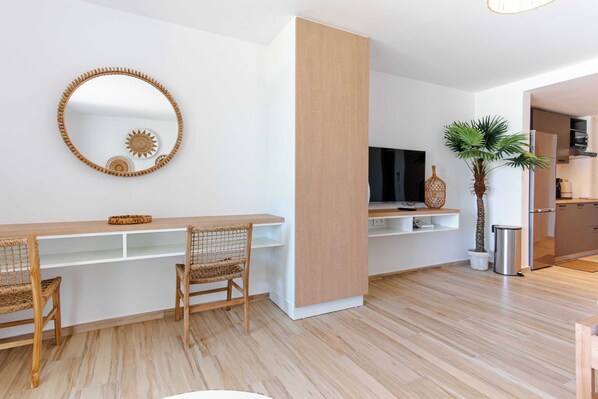 A modern living room with a wooden dining table, two wicker chairs, a large mirror, a flat-screen TV, a small plant, and a kitchen area with a refrigerator and microwave.