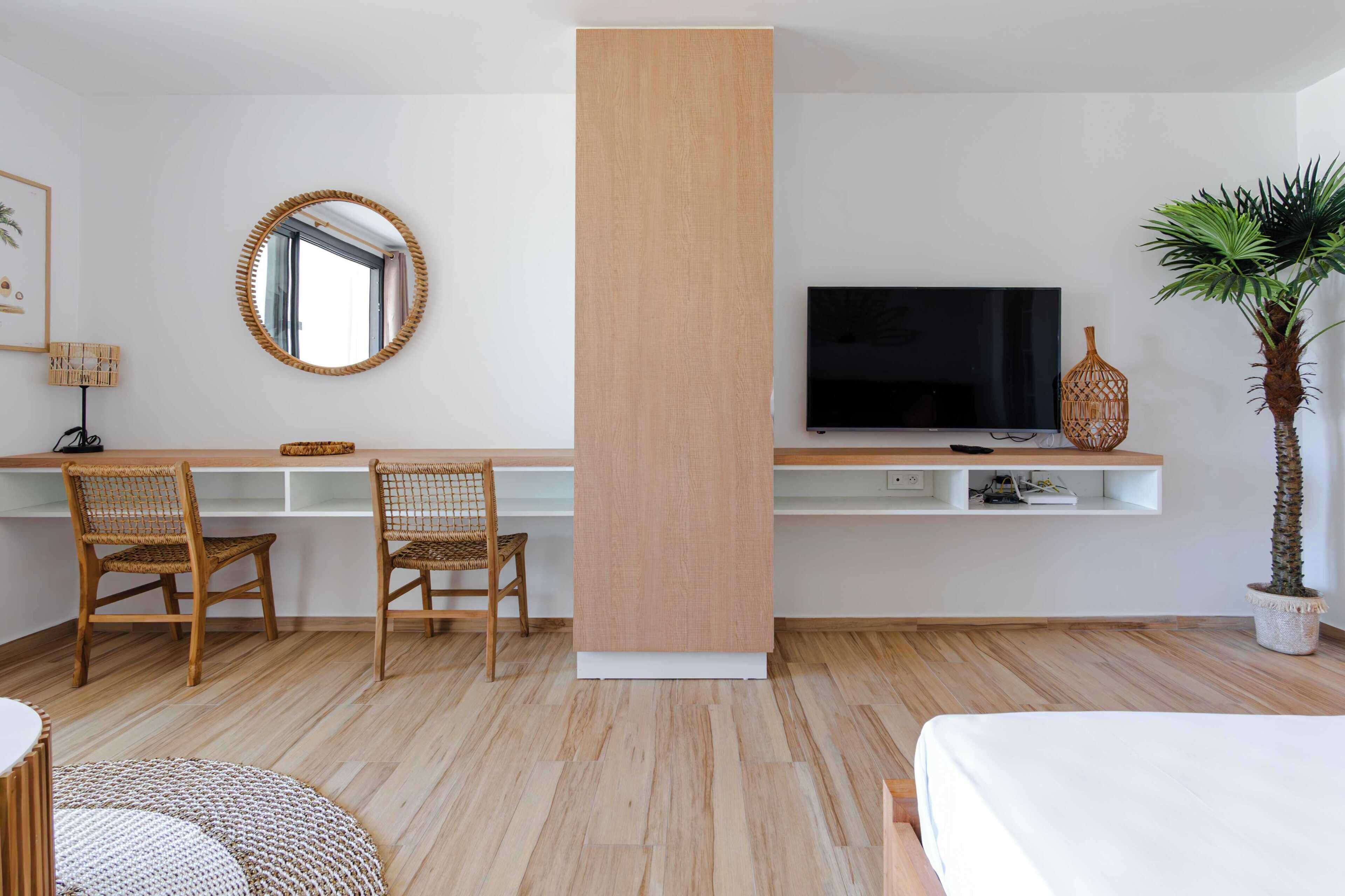 Une chambre d’hôtel moderne avec un bureau en bois, deux fauteuils en osier, un grand miroir, un téléviseur à écran plat, une plante en pot et un lit.