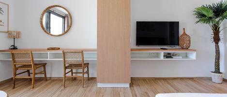 Une chambre d’hôtel moderne avec un bureau en bois, deux fauteuils en osier, un grand miroir, un téléviseur à écran plat, une plante en pot et un lit.