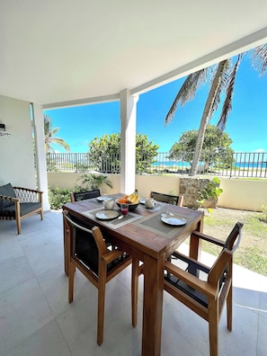 Mesa de madera con platos, cubiertos y un frutero, rodeada de sillas, con vista a una playa y palmeras.