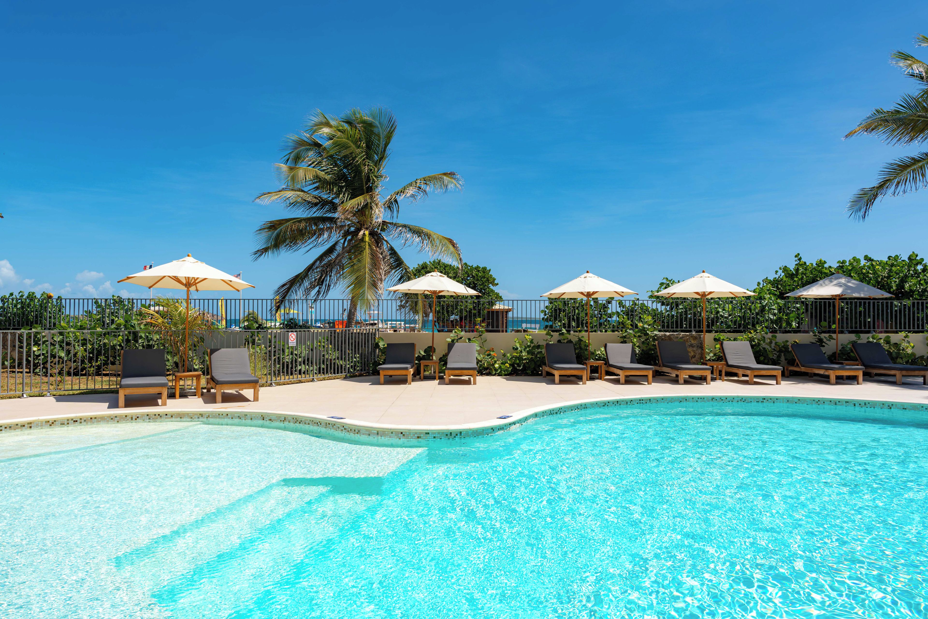 A swimming pool with lounge chairs and umbrellas, surrounded by a fence and palm trees.