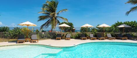 A swimming pool with lounge chairs and umbrellas, surrounded by a fence and palm trees.
