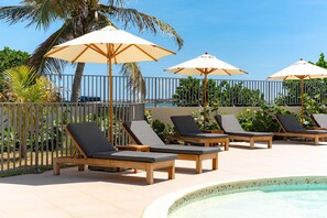 A poolside area with lounge chairs and umbrellas, surrounded by a fence and palm trees.