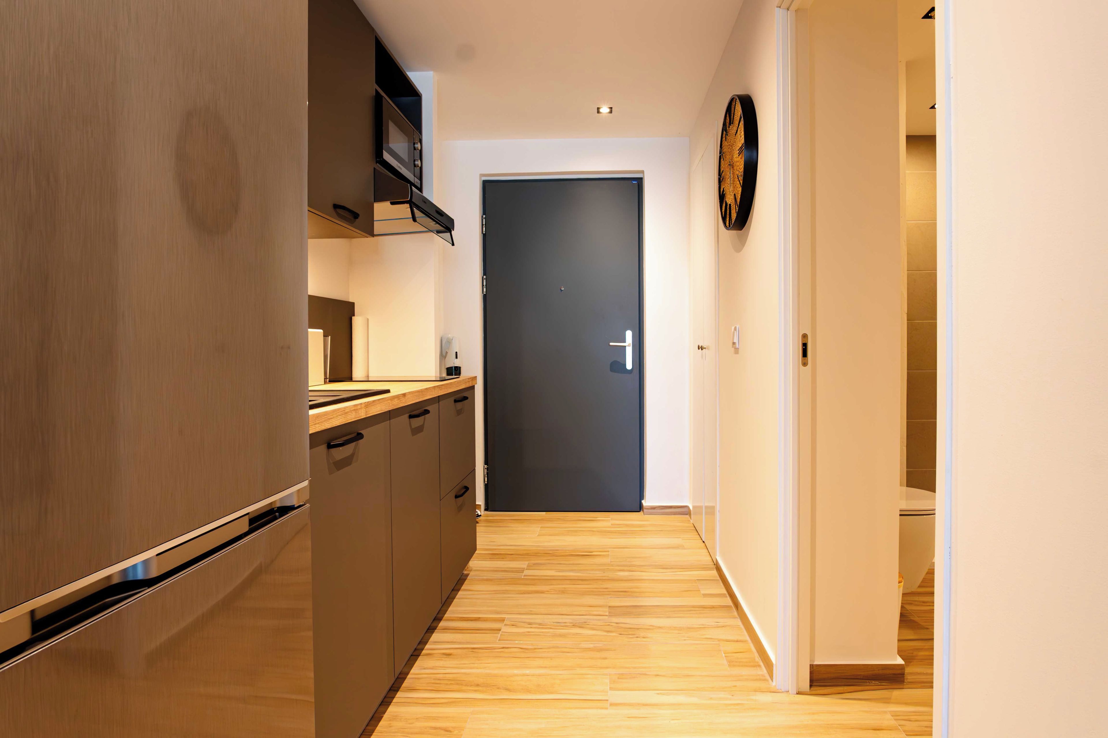A narrow hallway with wooden flooring leading to a dark door, flanked by kitchen cabinets and a wall-mounted clock.