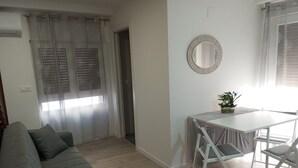 Een woonkamer met een grijze bank, een eettafel met stoelen, een kleine potplant en een ronde spiegel aan de muur.