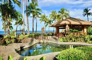 A tropical resort with a pool, palm trees, and a thatched-roof pavilion.