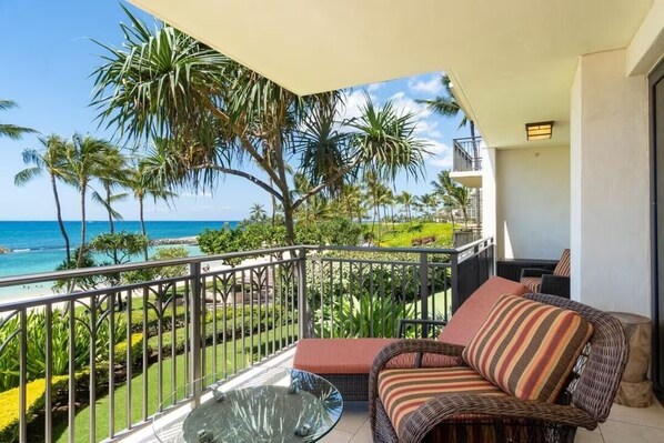 A balcony with a wicker sofa and a glass table, overlooking a beach with palm trees and the ocean.