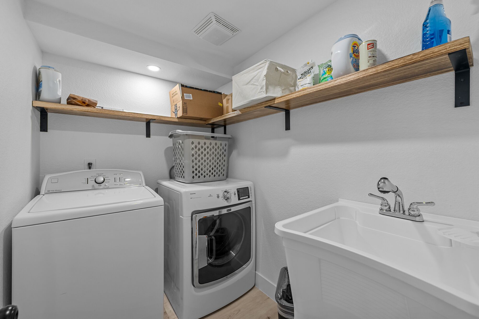 Een kleine wasruimte met een wasmachine, droger en wastafel. Daarboven zijn houten planken met schoonmaakmiddelen en een mand.