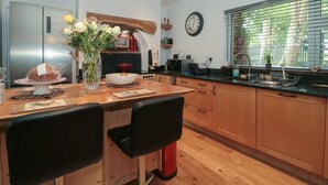 A modern kitchen with wooden cabinets, a stainless steel refrigerator, a dining area with two chairs, a vase with flowers, and a clock on the wall. - Brook Farm (Ringwood)