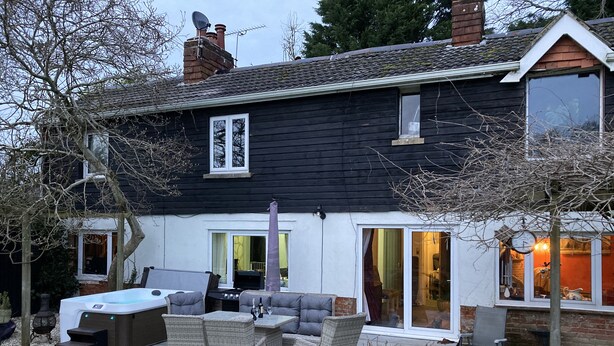 A modern two-story house with a patio area featuring outdoor furniture, a hot tub, and a large window overlooking the garden. - Brook Farm (Ringwood)