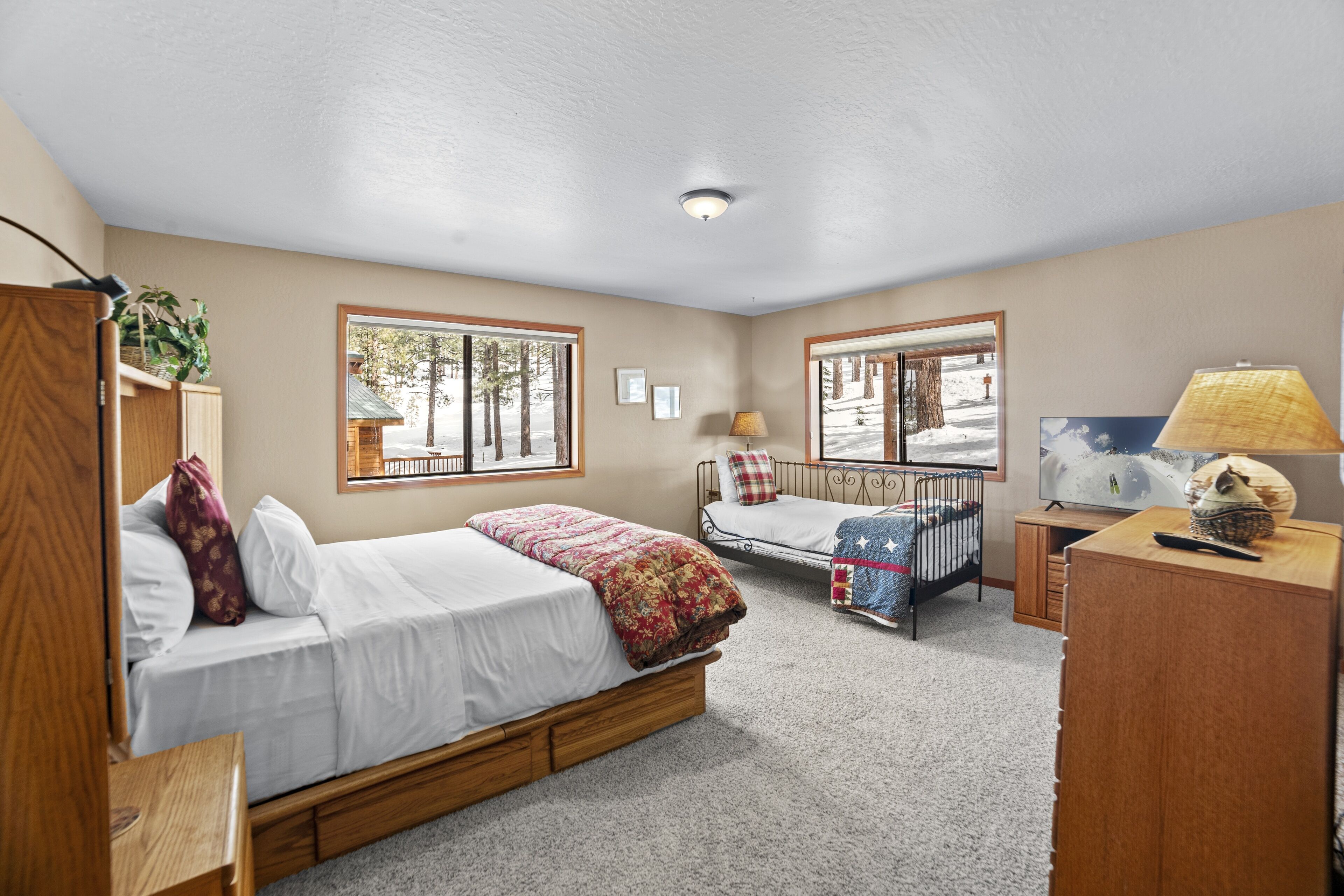 A bedroom with a large bed, a smaller bed, a desk, and a TV. There are windows and a ceiling light.