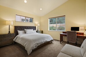 Un dormitorio con cama, escritorio, silla, ventana con vista a follaje y mesita de noche con lámpara.