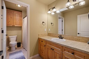 Un baño con inodoro blanco, armarios de madera, encimera color beige y un espejo con iluminación.