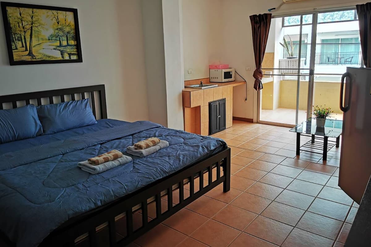 A bedroom with a bed, a small kitchenette, a balcony with a view, and a painting on the wall.