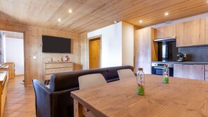 Een houten eettafel met een aan de muur bevestigde televisie, een keuken met kasten, en een zithoek met een bank.