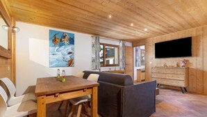 Een kamer met houten lambrisering, een eettafel, stoelen, een bank, een televisie en een schilderij aan de muur.