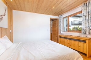 Een slaapkamer met een houten plafond, een bed met witte beddengoed, een houten dressoir en een raam met bloemen gordijnen.