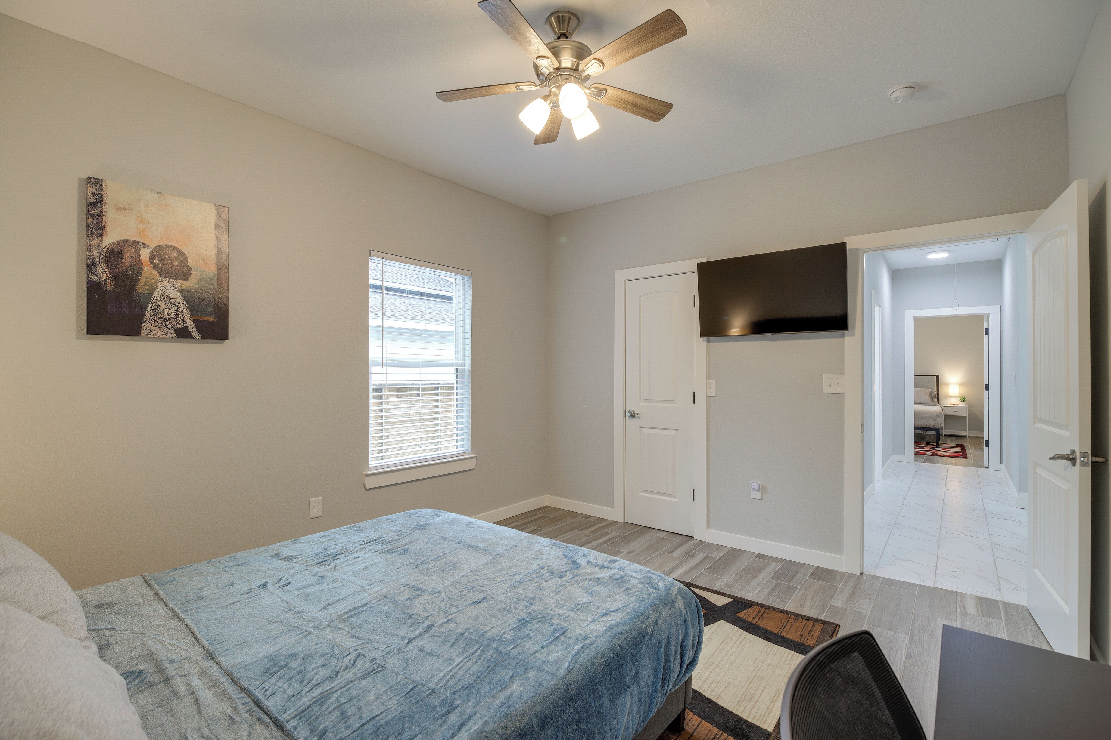 A bedroom with a bed, a ceiling fan, a painting, a window, and a door leading to another room.