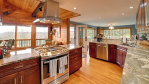 A spacious kitchen with wooden cabinets, granite countertops, stainless steel appliances, and large windows offering views of the outdoors. - Spacious Finger Lakes Home w/ Mountain Views (Dryden)