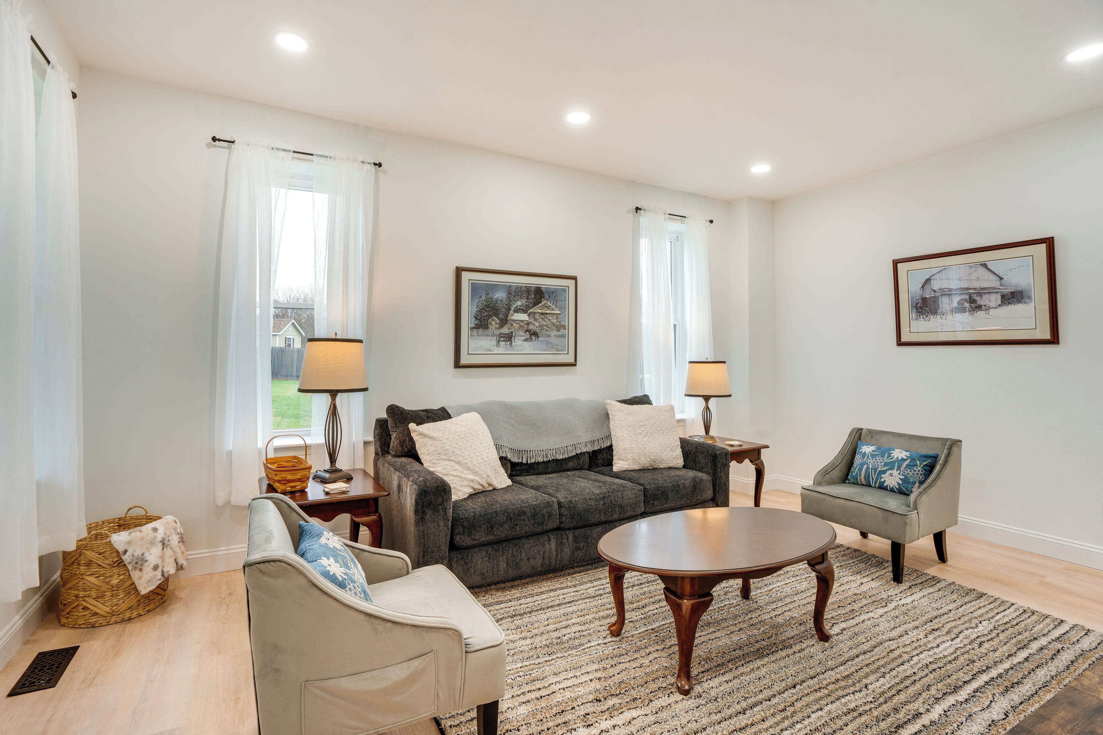 A living room with a grey sofa, a white armchair, a wooden coffee table, and a patterned rug. There are two lamps on side tables, a basket, and a framed picture on the wall.