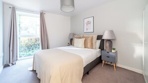 A bedroom with a bed, bedside tables, a lamp, a framed picture on the wall, and a window with curtains. - The Lewisham Collection (London)