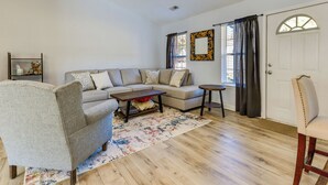 A living room with a sectional sofa, a small round table, and a chair. There is a wooden floor and a window with curtains.